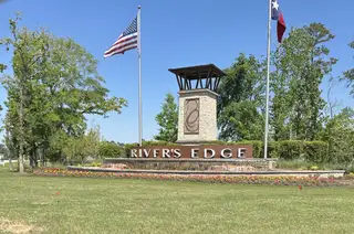 Street view A welcoming entrance sign for River's Edge by Legend Homes, adorned with flags and surrounded by verdant greenery in Conroe, TX.