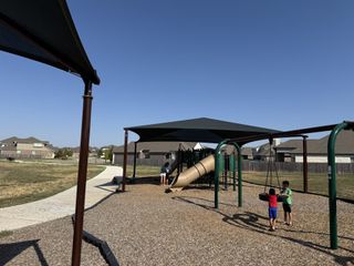 Community Amenities A fun playground with green slides and climbing structures, surrounded by shade trees in Blackhawk by GFO Home (Pflugerville, TX).