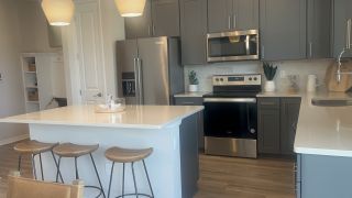 Model Home A modern kitchen with gray cabinets, stainless appliances, and a white island. Pendant lighting and wood floors add warmth.