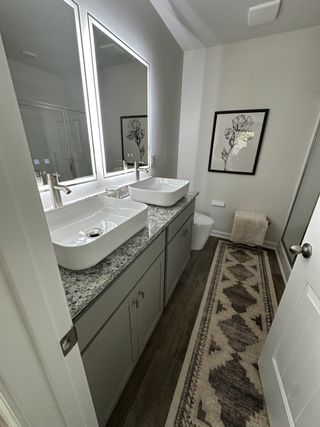 A sleek bathroom with dual vessel sinks, granite countertop, lit mirrors, and elegant decor on polished wood flooring.