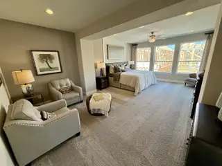 Model Home A spacious bedroom with plush carpet, neutral tones, and ample natural light. A sitting area adds comfort to the serene and inviting space.