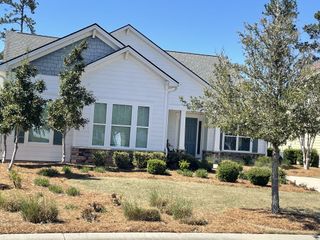 A bright, single-story home with white siding, blue accents, and lush landscaping, in Four Seasons at Lakes of Cane Bay by K. Hovnanian® Homes, Summerville, SC.