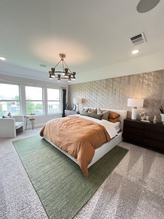 A spacious bedroom with a contemporary chandelier, textured accent wall, plush carpet, and large windows offering abundant natural light.