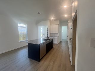 A bright, modern kitchen with a dark island, granite countertops, stainless steel appliances, and light wood flooring, leading to a hallway.
