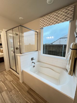 Model Home A modern bathroom featuring a sleek bathtub, glass shower, wood-look tiles, and large window with patterned valance.