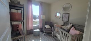 A charming nursery in a Marbella home, featuring a gray crib, a comfortable armchair, pink and gray curtains, and decorative mirrors (San Antonio, TX).