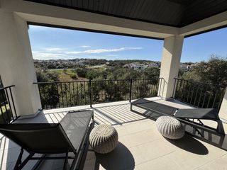 Model Home A sun-drenched balcony with lounge chairs and wicker poufs, offering a panoramic view of the surrounding hills and residential area.
