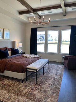 Model Home A cozy bedroom featuring a beam ceiling, elegant chandelier, and large windows with a view, complemented by rich textiles.