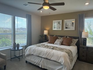 Model Home A cozy bedroom with a plush bed, decorative pillows, matching lamps, framed artwork, and a ceiling fan, creating a warm and inviting space.