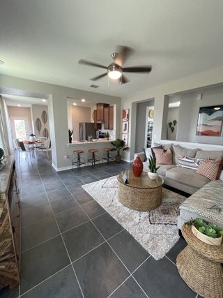 Model Home A spacious living area in a Mission del Lago home, featuring dark gray tile flooring, a comfortable gray sofa, a woven coffee table, and an open layout leading to the dining area and kitchen (San Antonio, TX).