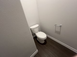 A clean, minimalist powder room features a white toilet, a chrome toilet paper holder, and dark wood-look flooring, offering a simple and functional space.