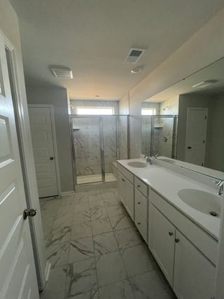 A spacious bathroom features a dual vanity with white cabinetry, a glass-enclosed shower with marble-look tiles, and large mirrors reflecting light.