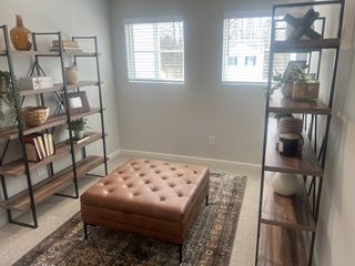 Model Home A versatile space features a tufted leather storage ottoman, flanked by two industrial-style bookshelves and illuminated by natural light from dual windows.