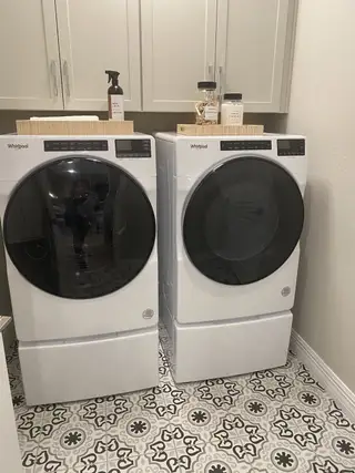 A functional laundry room features white Whirlpool appliances, light gray cabinetry with black hardware, wooden trays with decorative items, and patterned tile flooring.