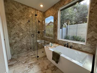 A luxurious bathroom with herringbone tiles, glass shower, and a deep soaking tub by a large window with garden views.