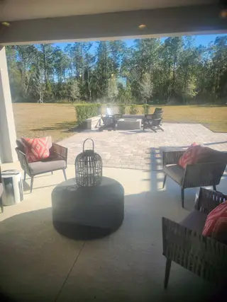 Model Home A cozy patio with wicker seating, patterned cushions, and a view of a serene backyard with a fire pit and lush trees.