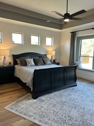 A chic bedroom with a sleek black bed, elegant lamps, wood floors, and a plush rug under a stylish ceiling fan.
