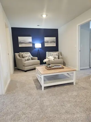 A cozy living area features plush carpeting, two armchairs with decorative pillows, a dark blue accent wall with framed artwork, and a light wood coffee table with a woven basket, illuminated by a modern floor lamp.