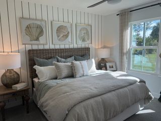 A cozy bedroom featuring a wicker headboard, nautical wall art, and plush bedding with natural light from a large window.