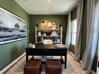 A cozy home office with green walls, stylish black desk, leather stools, and ample natural light from large windows.