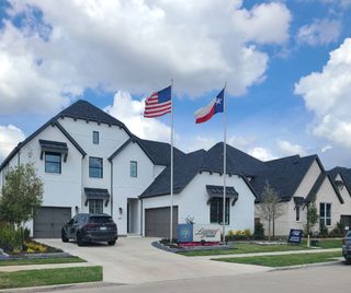 Street view Elegant white home with dark accents in The Grove Frisco - 65s by American Legend Homes, featuring flags and manicured landscaping.