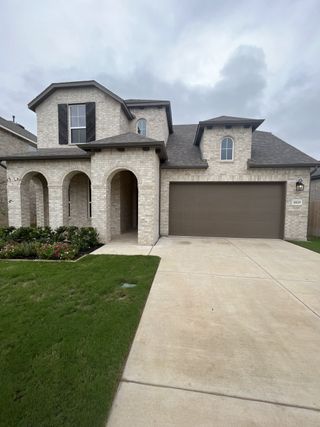 A stately light brick home in VIDA by Highland Homes (San Antonio, TX) features arched entryways, a dark garage door, and lush landscaping.