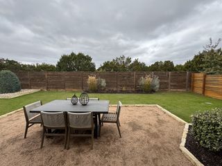A serene backyard with a dining set on gravel, surrounded by lush greenery and wooden fencing.