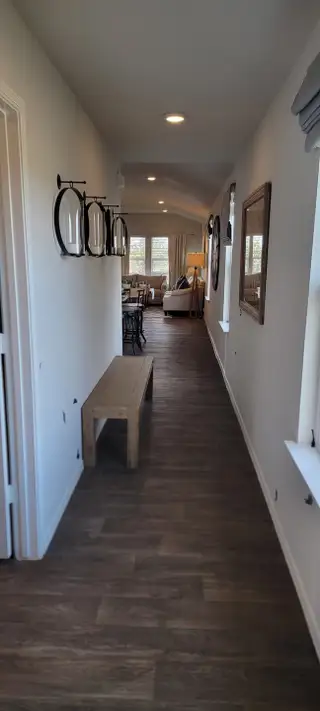 A long hallway with dark wood laminate flooring leads to a bright living area, featuring a simple wooden bench and decorative wall accents.