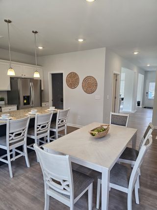 An open-concept living space features a modern kitchen with granite countertops and stainless steel appliances, a dining area with a light wood table and white chairs, and decorative woven wall hangings.
