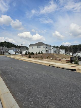 A peaceful suburban street showcases modern homes with light siding and dark roofs, nestled on gently rolling terrain under a bright, partly cloudy sky in Bailey Fence by Taylor Morrison (Dacula, GA).