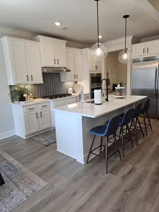 A bright kitchen with white cabinetry, a quartz-topped island with seating, pendant lighting, a stainless steel refrigerator, and a patterned backsplash.