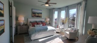 Model Home A serene bedroom with soft blue walls, a plush white bed, and a cozy sitting area by large windows, offering a tranquil retreat.