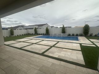 A beautifully landscaped backyard features a sparkling blue pool with a water feature, surrounded by a stylish stone patio with geometric grass inlays, and a tall privacy wall.