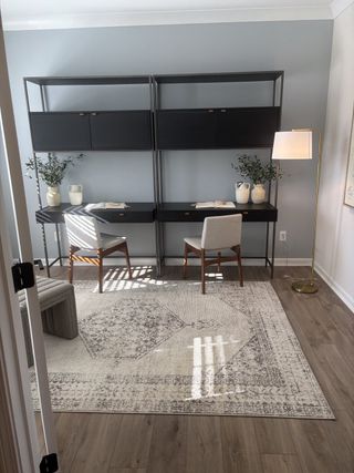 A chic home office with dual black desks, light gray walls, and a patterned rug, accented by modern chairs and decor.