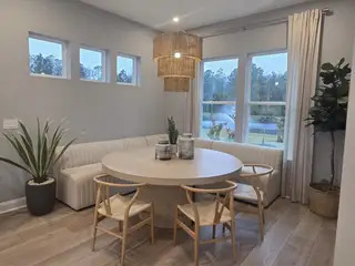 A cozy dining area featuring a round wooden table, stylish pendant lighting, and a plush banquette with large windows.