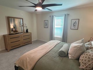 Model Home A cozy bedroom with a plush bed, wooden dresser, modern ceiling fan, and natural light filtering through soft curtains.