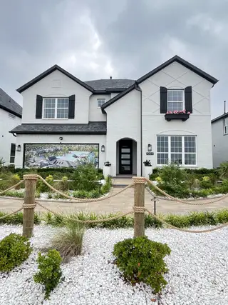 A charming white brick home with black shutters and a lush garden in Bellagio by Megatel Homes (Forney, TX).