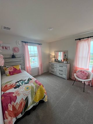 A cozy bedroom featuring floral bedding, soft pink accents, and natural light from two large windows.