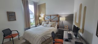 Cozy bedroom with a rustic headboard, neutral tones, a small desk, and natural light streaming through large windows.