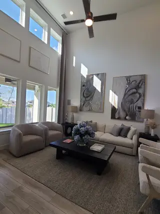A cozy living room with high ceilings, large windows, abstract art, neutral furniture, and a modern ceiling fan.