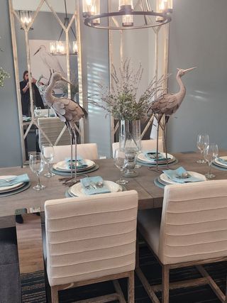 A chic dining room featuring elegant mirrors, bird sculptures, soft lighting, and a tastefully set table with plush seating.