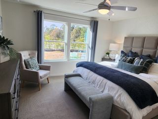 A serene bedroom featuring a cozy upholstered bed, soft carpeting, modern ceiling fan, and plentiful natural light from large windows.