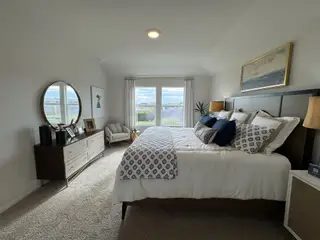 A serene primary suite with plush carpeting, a spacious bed, and large windows offering backyard views.