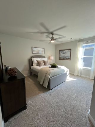 A cozy bedroom featuring a plush bed, soft carpeting, modern ceiling fan, and large window with soft natural light.