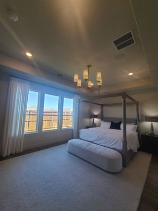 A cozy bedroom with a four-poster bed, large windows, elegant chandelier, and soft carpeting for a relaxing atmosphere.