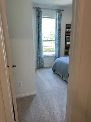 A cozy bedroom with soft carpet, blue bedding, and patterned curtains framing a bright window.