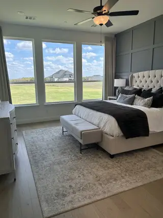 A serene bedroom with a plush bed, gray accent wall, large windows, and elegant decor on hardwood floors.