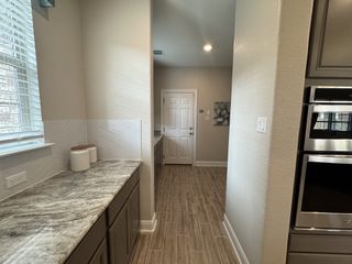 Model Home A narrow hallway with gray granite countertops, light wood-look tile flooring, and a white door at the end, leading to another room.