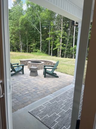 Model Home A cozy patio featuring a stone fire pit, paver flooring, and green Adirondack chairs overlooking a serene wooded area.