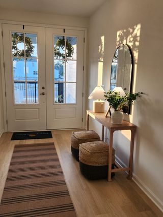 A welcoming entryway featuring double glass doors, a wooden console with decor, and cozy poufs, bathed in warm sunlight.